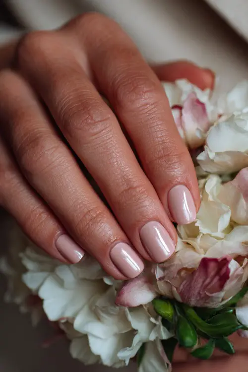 Hand of bridesmaid resting on floral corsage, classic rounded nails in soft pink-beige with a satin finish and delicate pearl dot at the base 