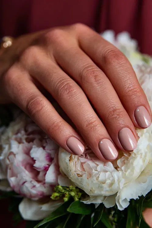 Hand of bridesmaid resting on floral corsage, classic rounded nails in soft pink-beige with a satin finish and delicate pearl dot at the base 