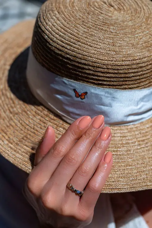 Hand of guest adjusting sun hat ribbon, short ballerina nails painted in pastel peach with sheer gloss and one mini butterfly decal