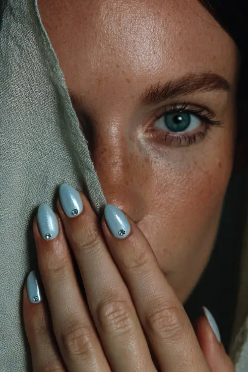 Hand tucking veil behind ear, almond nails in soft pastel blue with chrome shimmer and a single crystal stud at the base of each nail 