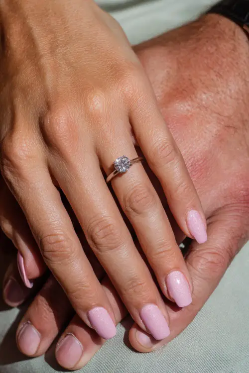 Soft close-up of interlocked bridal hands, wearing an engagement ring, with pastel pink nails and silver glitter accents