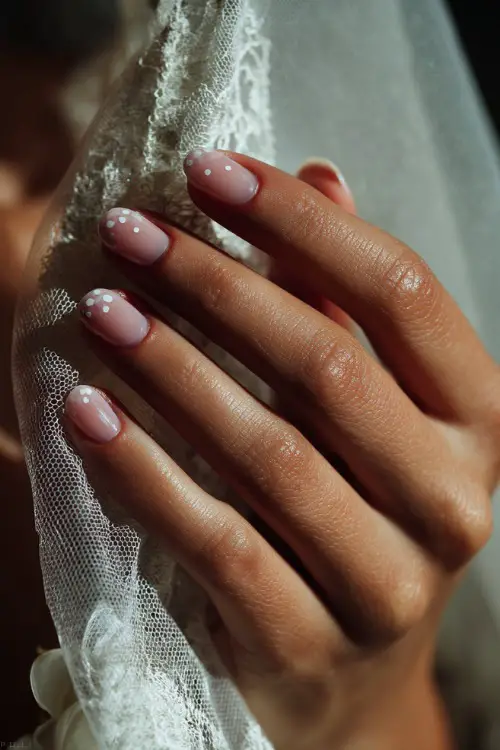 Soft light on a bride’s hand reaching for her veil, round nails in matte pale pink with delicate white dots and minimalistic flower petals on the tips 