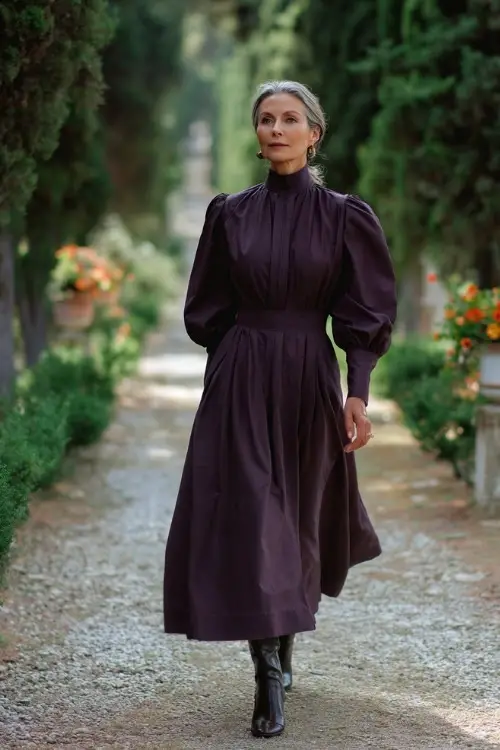 Elegant older woman in a high-neck dark plum midi dress with bishop sleeves and ankle boots, walking along a tree-lined garden aisle