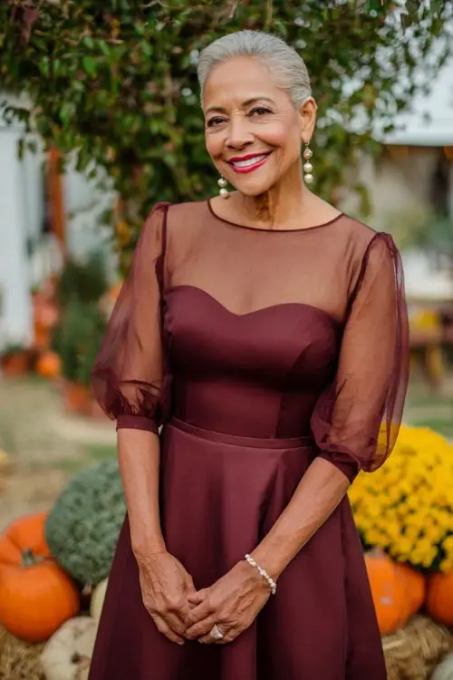 Elegant older woman wearing a burgundy fit-and-flare midi dress with sheer sleeves and pearl earrings, standing near pumpkins and mums at a fall wedding setting 