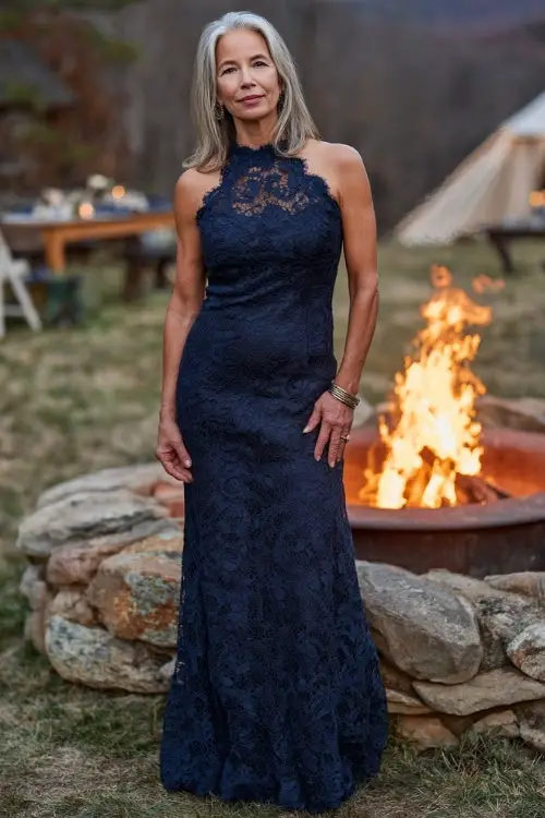 Elegant woman over 50 in a navy lace gown with scalloped edges and high neckline, standing near a glowing outdoor fire pit at a fall wedding reception 