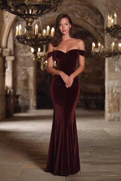 Elegant woman wearing a burgundy velvet off-shoulder mermaid dress with a subtle train, standing inside a candlelit stone hall with vintage chandeliers 