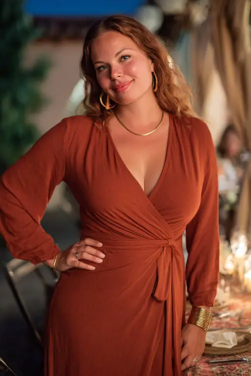 Full body of a curvy woman in a rust-colored midi wrap dress with long sleeves and gold accessories, posing at a candlelit outdoor wedding in early evening light