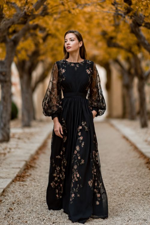 Full body of a woman in a black floor-length gown with sheer long sleeves and floral embroidery, posing under a canopy of amber maple trees at a luxury outdoor wedding