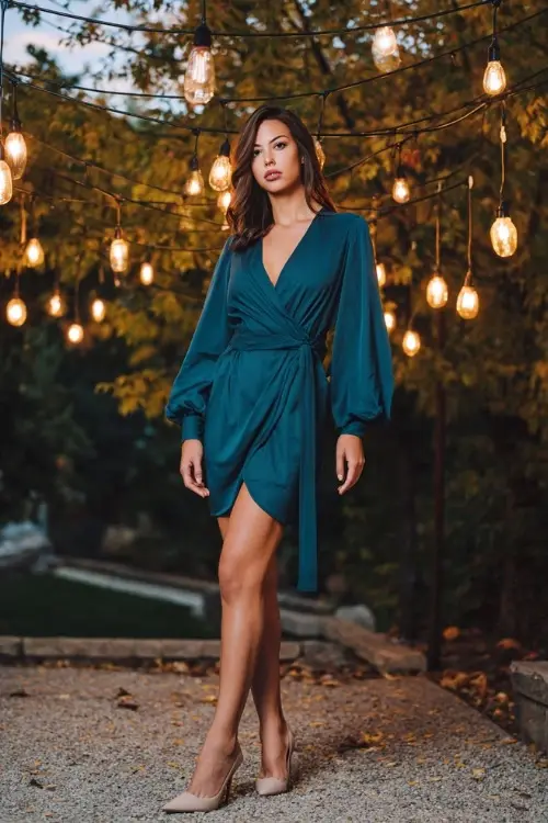 Full body of a woman in a dark teal wrap dress with balloon sleeves and pointed heels, standing under hanging lights with early fall trees in the background