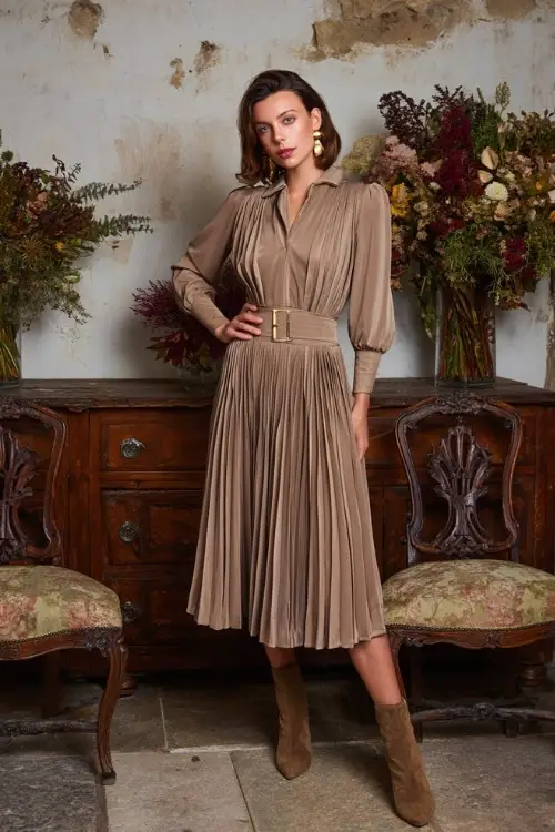 Full body of a woman in a taupe pleated dress with long sleeves and high waist, styled with suede ankle boots and gold earrings, standing beside vintage chairs and seasonal floral décor 