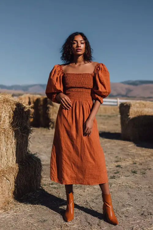 Full body of a woman wearing a burnt orange midi dress with puff sleeves and smocked bodice, styled with ankle boots at a backyard wedding with hay bales and pumpkins
