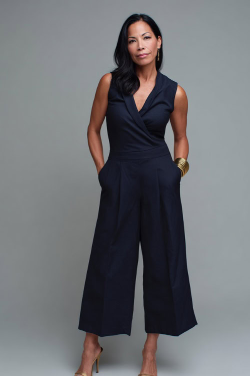 Mature elegant woman wearing a navy wide-leg jumpsuit with a wrap front, gold cuff bracelet, and heeled mules, in a clean and soft-lit studio
