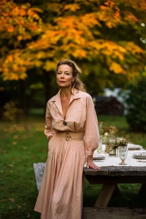 Mature woman in a blush long sleeve midi dress with flowy skirt and brooch detail, posing near a rustic table setup under golden maple trees 