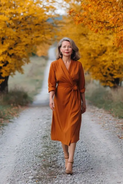 Mature woman in a cinnamon-colored midi dress with soft draping and long sleeves, walking on a gravel path framed by golden autumn trees