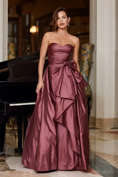 Mature woman in a dusty rose formal gown with satin bow waist and structured silhouette, posing near a piano in a high-end fall wedding venue