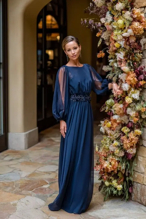 Mature woman wearing a navy blue chiffon gown with sheer sleeves and embellished waist, posing beside a fall floral arch at an upscale wedding 