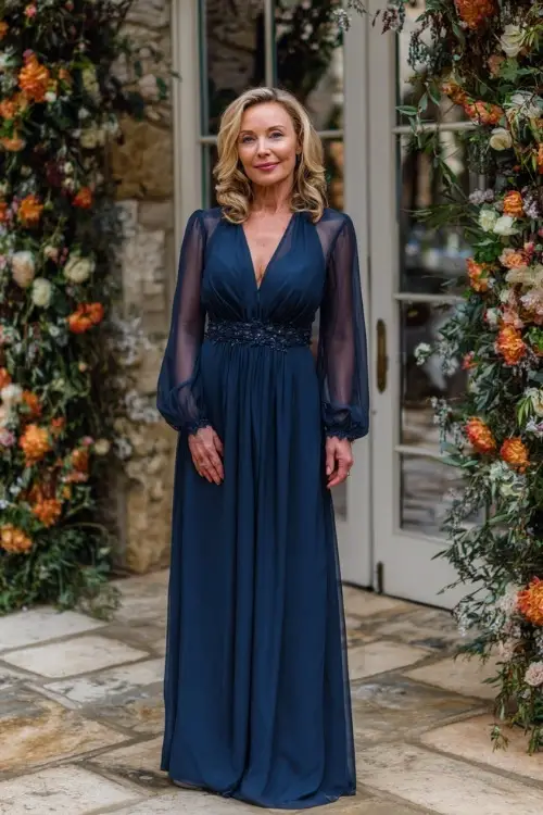 Mature woman wearing a navy blue chiffon gown with sheer sleeves and embellished waist, posing beside a fall floral arch at an upscale wedding 