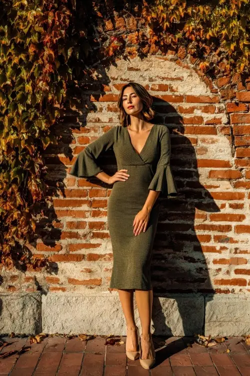 Mature woman wearing an olive green midi dress with subtle shimmer and flared sleeves, posing by a vintage brick wall draped in fall ivy, full body