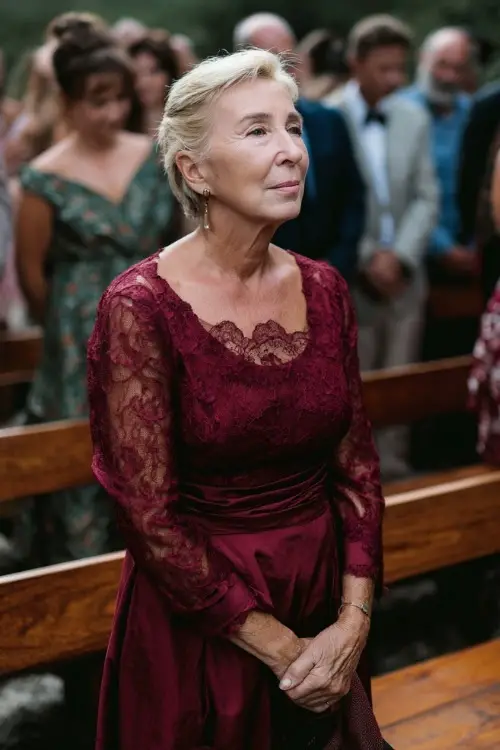 Older woman in a maroon midi dress with lace overlay and long sleeves, standing at an outdoor wedding ceremony with wooden benches, full body visible