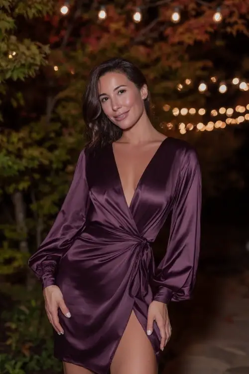 Short height woman wearing a deep aubergine satin wrap dress with long balloon sleeves and a subtle slit, posing under string lights and warm-toned fall foliage