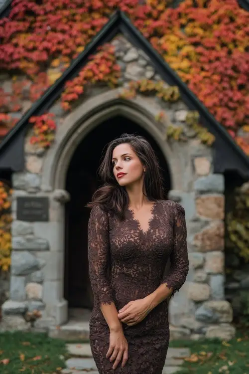 Woman in a chocolate lace dress with fitted silhouette and mesh long sleeves, standing in front of an old stone chapel covered in colorful fall vines