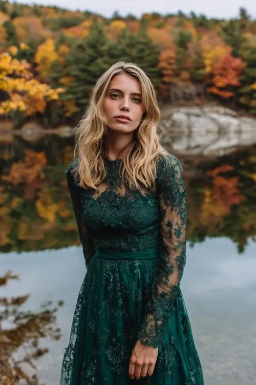 Woman in a forest green midi dress with lace overlay and long sheer sleeves, standing at a lakeside wedding with fall foliage reflections, full body visible
