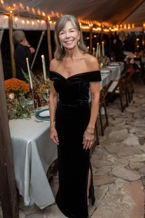 Woman over 50 in a black velvet off-shoulder gown with a side slit and pearl earrings, standing beside an evening fall wedding reception setup with candlelight 