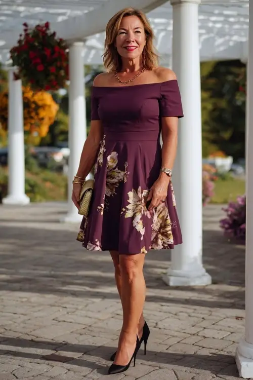 Woman over 50 in a plum-colored fit-and-flare dress with boat neckline and gold accessories, standing near a white pergola decorated with September fall florals 