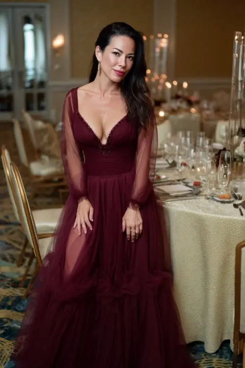 Woman over 50 in a rich burgundy gown with flowing tulle skirt and embellished neckline, posing beside elegant banquet tables set for a fall wedding