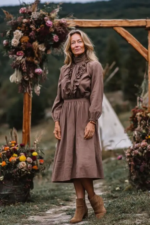 Woman over 50 in a soft brown high-neck midi dress with ruffle trim and tan ankle boots, posing near a wooden ceremony arch decorated with early autumn flowers 