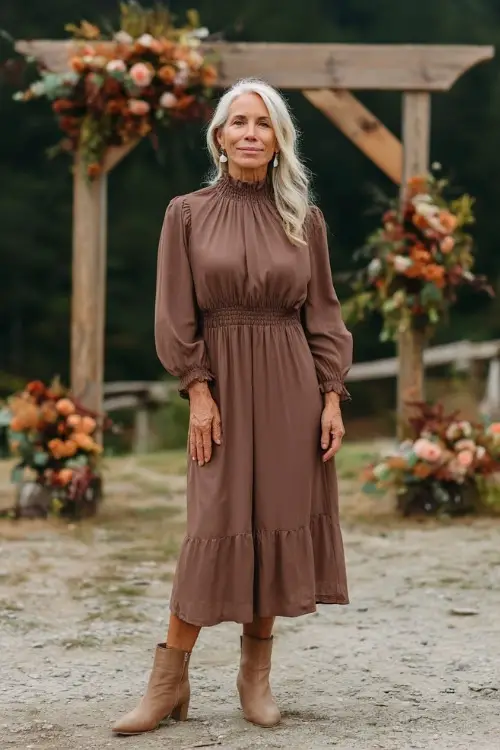 Woman over 50 in a soft brown high-neck midi dress with ruffle trim and tan ankle boots, posing near a wooden ceremony arch decorated with early autumn flowers