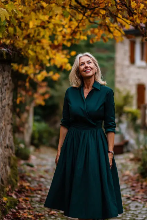 Woman over 50 wearing a deep forest green midi dress with long sleeves and pleated skirt, standing on a cobblestone path surrounded by fall leaves