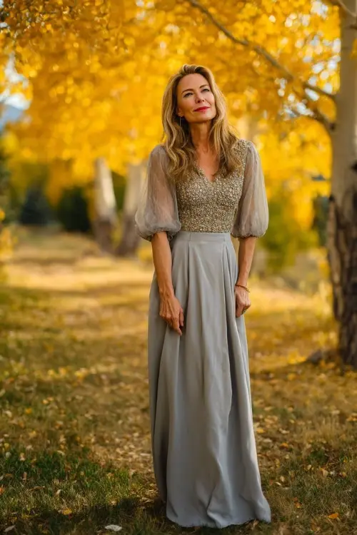 Woman over 50 wearing a silver-grey floor-length dress with beaded bodice and sheer sleeves, standing under a canopy of golden trees at sunset 