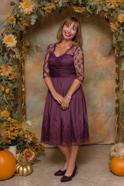 Woman over 50 wearing a tea-length plum dress with lace sleeves and classic black pumps, smiling near a floral arch with pumpkins and gold accents 