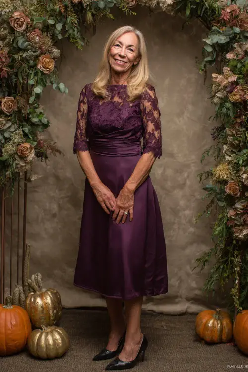 Woman over 50 wearing a tea-length plum dress with lace sleeves and classic black pumps, smiling near a floral arch with pumpkins and gold accents 