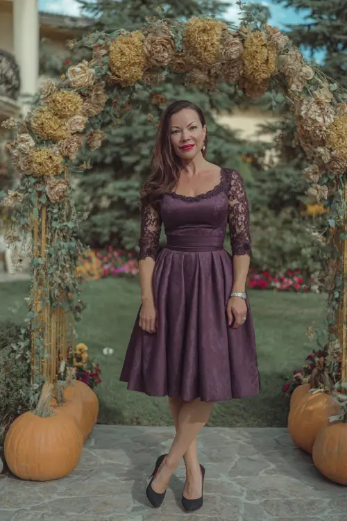 Woman over 50 wearing a tea-length plum dress with lace sleeves and classic black pumps, smiling near a floral arch with pumpkins and gold accents 