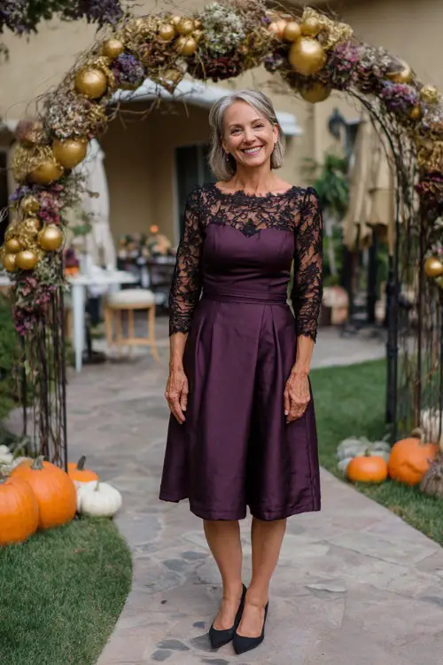 Woman over 50 wearing a tea-length plum dress with lace sleeves and classic black pumps, smiling near a floral arch with pumpkins and gold accents
