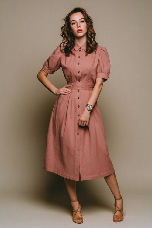 Woman wearing a muted rose tea-length dress with buttons down the front, round collar, tan heeled sandals, and vintage watch, minimalist studio background 