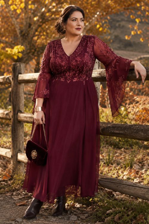 a woman wearing a burgundy dress with an embroidered fitted bodice, sheer bell sleeves, and a flowing ankle-length skirt
