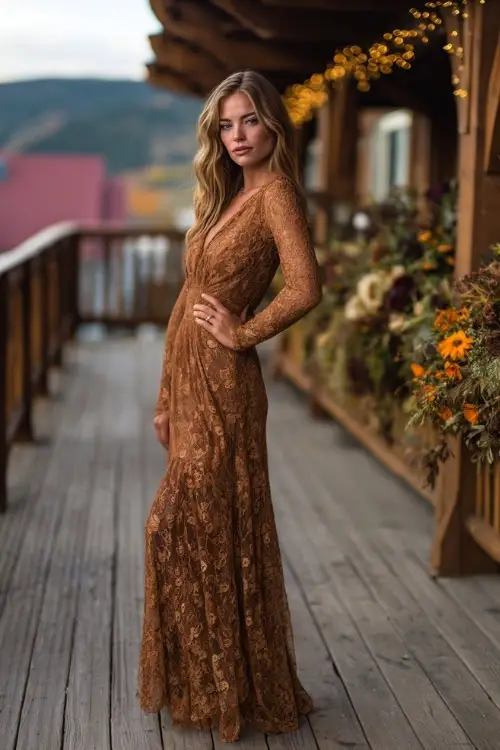 Full body of a woman in a copper-toned lace dress with long sleeves and flared hem, posing on a wooden deck decorated with fall florals and golden fairy lights