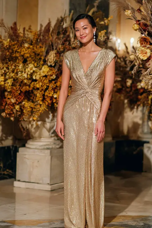 A woman in a champagne sequin gown with cap sleeves and a flowing skirt, accessorized with crystal earrings, standing in a ballroom decorated with golden fall florals 