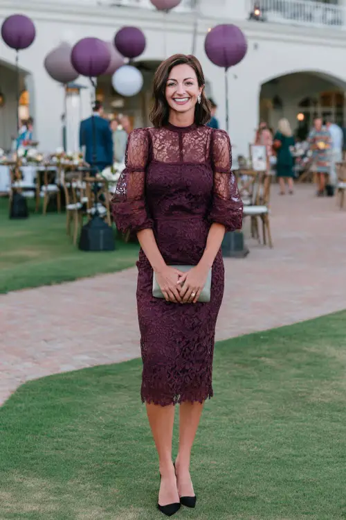 A woman in a deep plum lace midi dress with sheer sleeves, accessorized with a clutch and classic pumps, standing at a country club garden wedding reception decorated with lanterns 