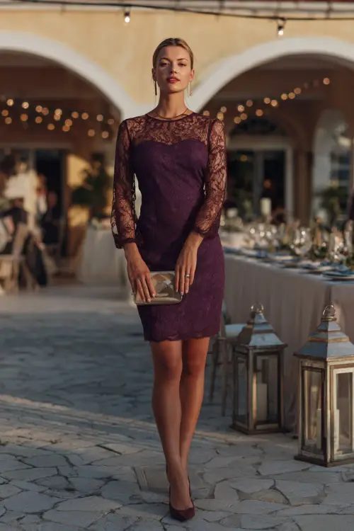 A woman in a deep plum lace midi dress with sheer sleeves, accessorized with a clutch and classic pumps, standing at a country club garden wedding reception decorated with lanterns