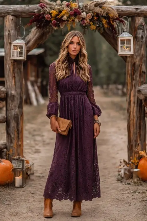 A woman in a plum lace maxi dress with long sleeves, styled with tan cowboy boots and a suede clutch, standing under a wooden arch decorated with fall florals and lanterns 