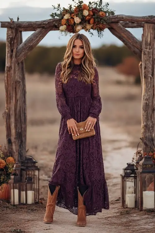 A woman in a plum lace maxi dress with long sleeves, styled with tan cowboy boots and a suede clutch, standing under a wooden arch decorated with fall florals and lanterns