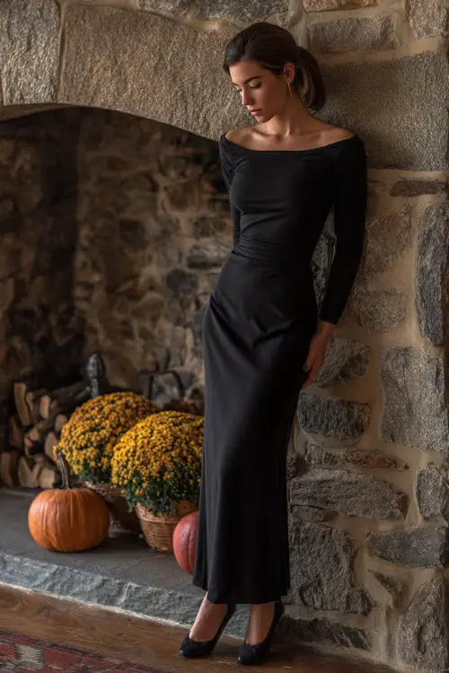 A woman in a simple long-sleeve black gown with a bateau neckline and floor-length skirt, paired with black pumps, standing near a stone fireplace with autumn décor 