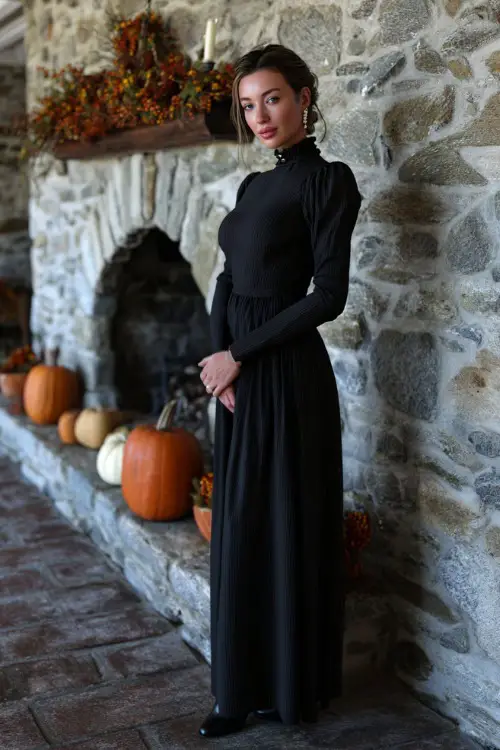 A woman wears a black crepe gown with long sleeves and high neckline, styled with pearl earrings and black pumps, standing near a stone fireplace decorated with pumpkins and fall garlands
