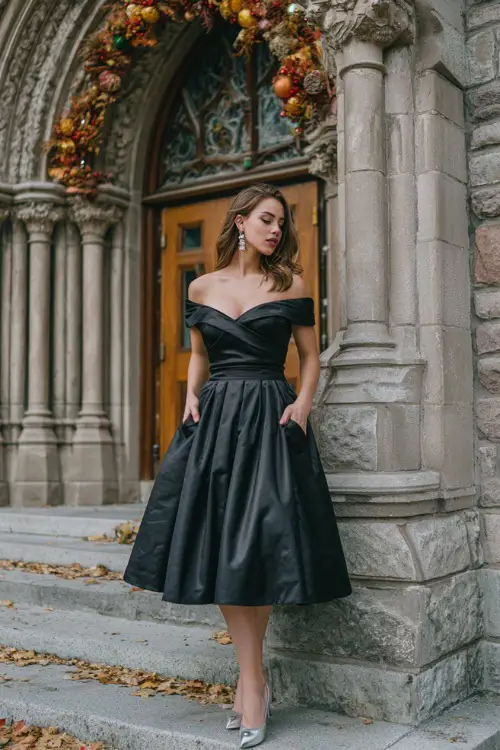 A woman wears a black off-the-shoulder ball gown with a voluminous skirt, styled with diamond studs and silver heels, standing in front of a historic cathedral entrance decorated for fall 