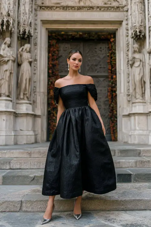 A woman wears a black off-the-shoulder ball gown with a voluminous skirt, styled with diamond studs and silver heels, standing in front of a historic cathedral entrance decorated for fall