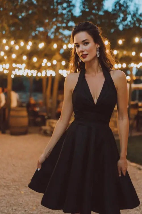 A woman wears a black short halter dress with a flared skirt, styled with silver earrings and classic pumps